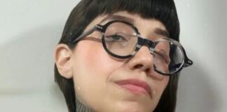 Dahlia de la Cerda in a publicity photo. The photo is a closeup of her face. She wears thick round black glasses and has long black hair and bangs. Some of her tattoos on her neck are visible.