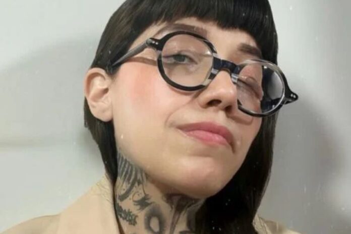 Dahlia de la Cerda in a publicity photo. The photo is a closeup of her face. She wears thick round black glasses and has long black hair and bangs. Some of her tattoos on her neck are visible.