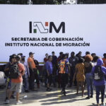 A crowd of migrants with backpacks wait in front of a wall reading "INM Secretaría de Gobernación," representing the reason why Mexico had to increase the INM budget