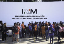 A crowd of migrants with backpacks wait in front of a wall reading "INM Secretaría de Gobernación," representing the reason why Mexico had to increase the INM budget