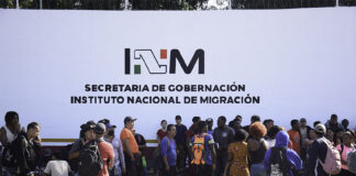 A crowd of migrants with backpacks wait in front of a wall reading "INM Secretaría de Gobernación," representing the reason why Mexico had to increase the INM budget