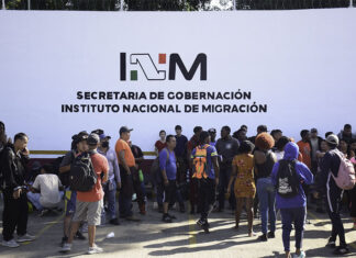 A crowd of migrants with backpacks wait in front of a wall reading "INM Secretaría de Gobernación," representing the reason why Mexico had to increase the INM budget