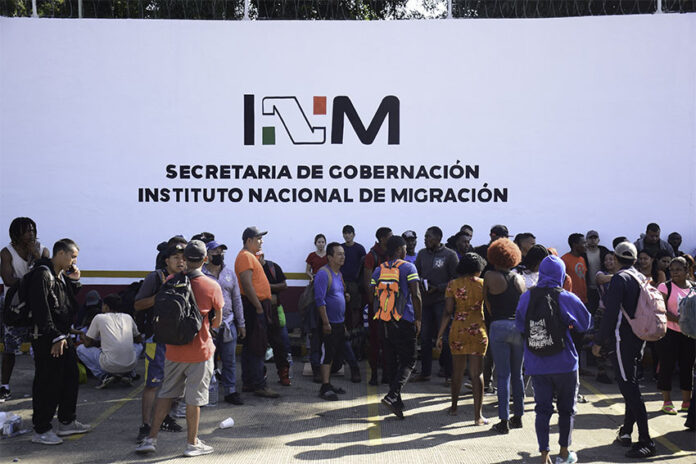 A crowd of migrants with backpacks wait in front of a wall reading 