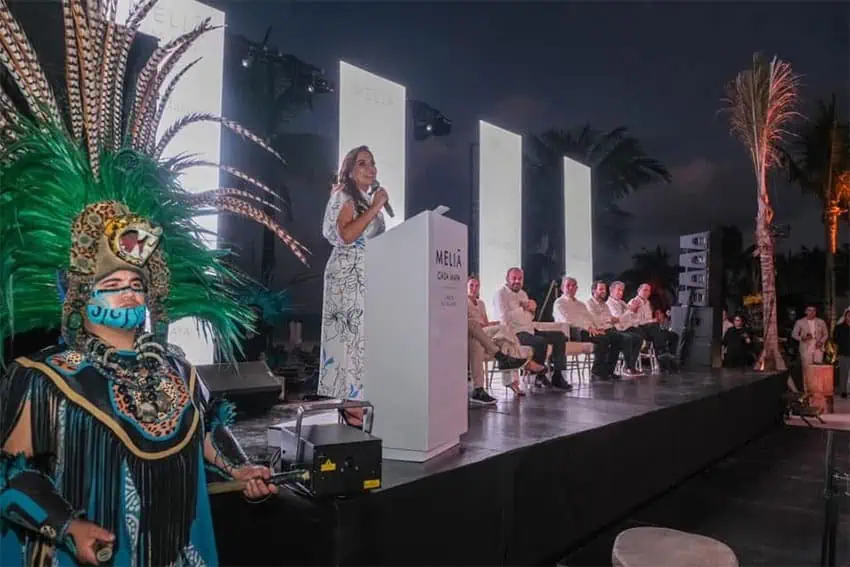 Quintana Roo Gov. Mara Lezama speaks into a microphone on a stage at night with dancers in feathered headdresses in the foreground