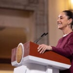 President Sheinbaum stands at a podium smiling during her Monday morning press conference, or mañanera