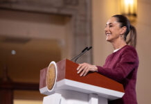 President Sheinbaum stands at a podium smiling during her Monday morning press conference, or mañanera