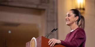 President Sheinbaum stands at a podium smiling during her Monday morning press conference, or mañanera