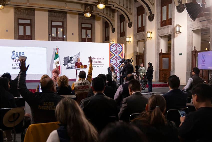 Mexican reporters in the foreground of the photo raising their hands to try and ask questions of President Claudia Sheinbaum at her daily press conference. Sheinbaum is standing at the presidential podium in the photo's distant background.