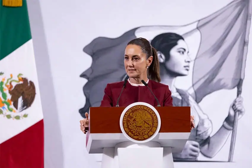 Mexican President Claudia Sheinbaum at the presidential podium during a press conference. She's wearing a burgundy blazer and holding onto the podium with her hands as she gazes at reporters off camera.