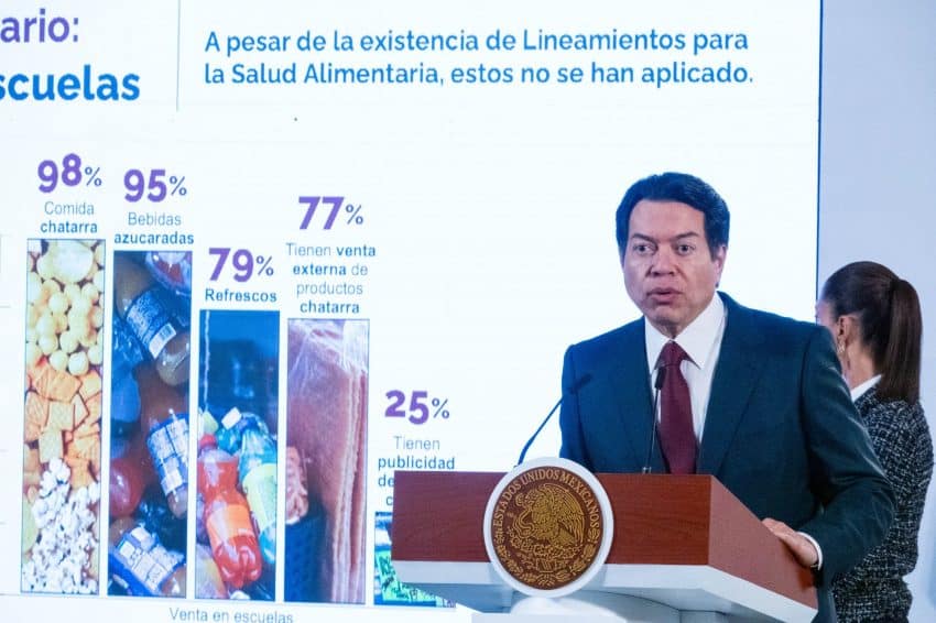 Un hombre con traje y corbata se encuentra en el podio presidencial en el Palacio Nacional de la Ciudad de México que habla con la prensa. Detrás de él hay una diapositiva de PowerPoint con gráficos que explican la cantidad de comida chatarra que se encuentra en las escuelas de México.