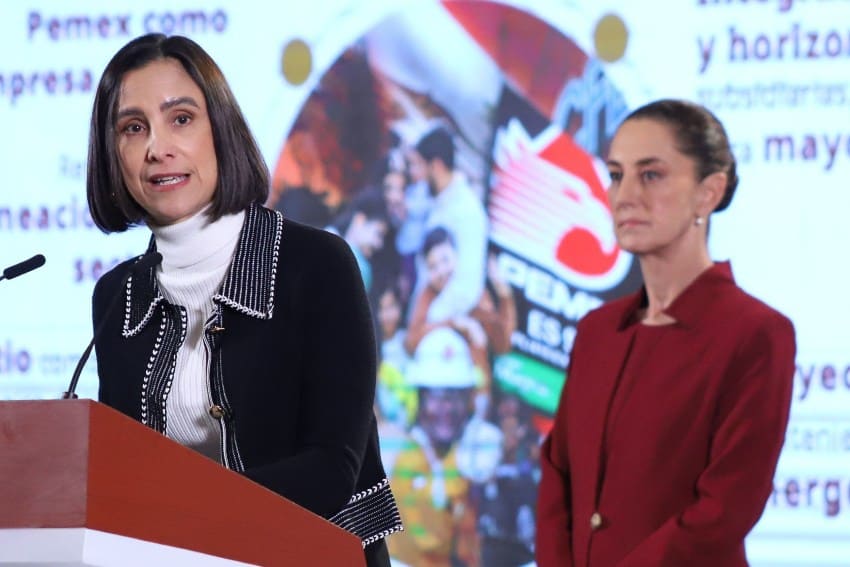 Mexico's Energy Minister Luz Elena Gonzalez standing at the presidential podium, talking to reporters at President Sheinbaum's daily press conference. Behind her, listening, stands President Sheinbaum.