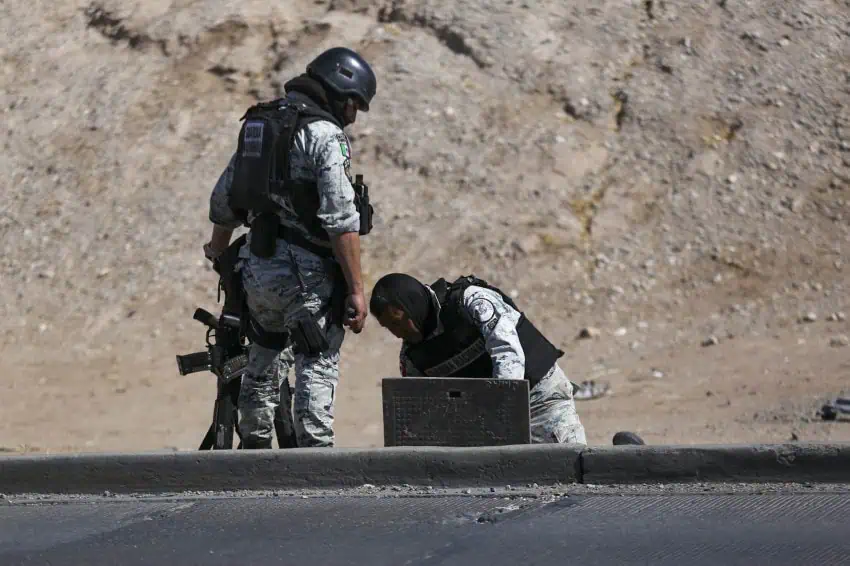 Dos guardias nacionales mexicanos que inspeccionan un presunto túnel ilegal construido para la trata de la frontera de México-EE. UU. Uno está parado sobre el túnel, que tiene una cubierta de hierro. El otro está arrodillado y revisando algo sobre el túnel.