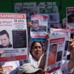 Mexican men and women at a protest in Mexico City, holding up laminated missing persons posters of their loved ones. All the posters say at the top "Desaparecido" (Disappeared/Missing)