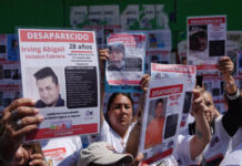 Mexican men and women at a protest in Mexico City, holding up laminated missing persons posters of their loved ones. All the posters say at the top "Desaparecido" (Disappeared/Missing)