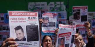 Mexican men and women at a protest in Mexico City, holding up laminated missing persons posters of their loved ones. All the posters say at the top "Desaparecido" (Disappeared/Missing)