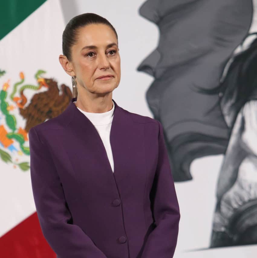 President Claudia Sheinbaum standing at a press conference in front of a wall at the National Palace with an image of the Mexican flag.