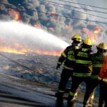 State firefighters at a raging fire in Monterrey. We see the backs of four firefighters in uniform and ahead of them, there is a wall of smoke clouds and downed electrical wires on the street.