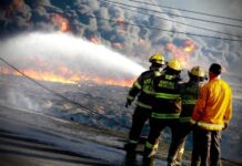 State firefighters at a raging fire in Monterrey. We see the backs of four firefighters in uniform and ahead of them, there is a wall of smoke clouds and downed electrical wires on the street.