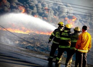 State firefighters at a raging fire in Monterrey. We see the backs of four firefighters in uniform and ahead of them, there is a wall of smoke clouds and downed electrical wires on the street.