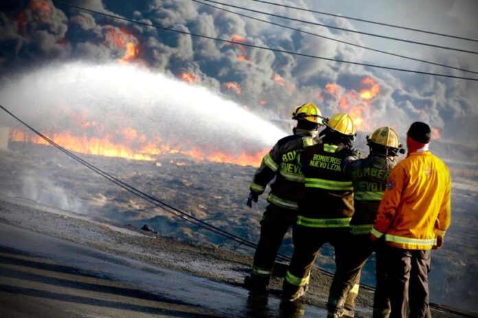 State firefighters at a raging fire in Monterrey. We see the backs of four firefighters in uniform and ahead of them, there is a wall of smoke clouds and downed electrical wires on the street.