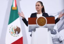 President Claudia Sheinbaum of Mexico standing at the presidential podium during her daily press conference. She is talking to reporters with her hands stretched out on either side of her body.