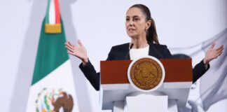 President Claudia Sheinbaum of Mexico standing at the presidential podium during her daily press conference. She is talking to reporters with her hands stretched out on either side of her body.