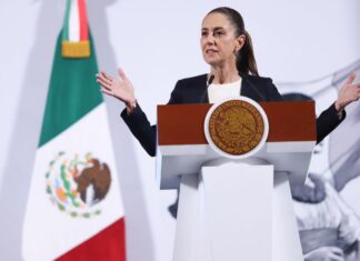 President Claudia Sheinbaum of Mexico standing at the presidential podium during her daily press conference. She is talking to reporters with her hands stretched out on either side of her body.