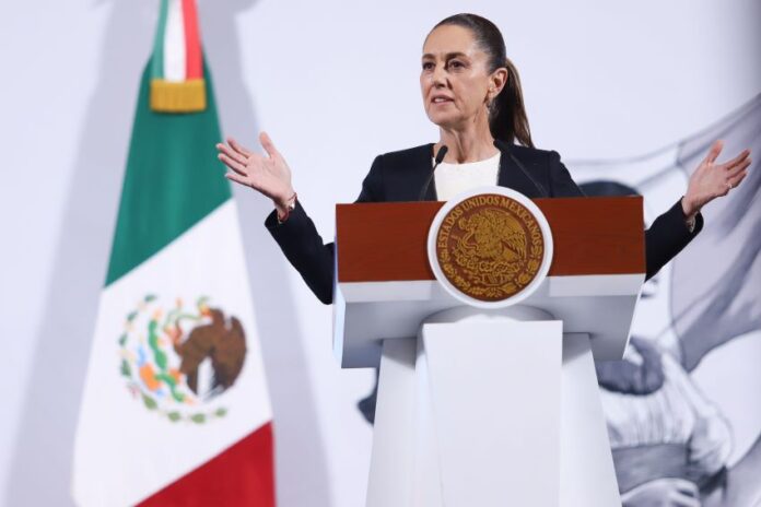 President Claudia Sheinbaum of Mexico standing at the presidential podium during her daily press conference. She is talking to reporters with her hands stretched out on either side of her body.