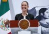 President Claudia Sheinbaum standing at the presidential podium at the National Palace with a wide smile as she gazes out at reporters off camera. Behind her is a large Mexican flag on a post and a wall with the logo of Sheinbaum's admnistration: an illustration of a young Indigenous Mexican woman in side profile looking off into the distance, standing in front of the Mexican flag.