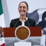 President Claudia Sheinbaum standing at the presidential podium at the National Palace with a wide smile as she gazes out at reporters off camera. Behind her is a large Mexican flag on a post and a wall with the logo of Sheinbaum's admnistration: an illustration of a young Indigenous Mexican woman in side profile looking off into the distance, standing in front of the Mexican flag.