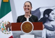 President Claudia Sheinbaum standing at the presidential podium at the National Palace with a wide smile as she gazes out at reporters off camera. Behind her is a large Mexican flag on a post and a wall with the logo of Sheinbaum's admnistration: an illustration of a young Indigenous Mexican woman in side profile looking off into the distance, standing in front of the Mexican flag.