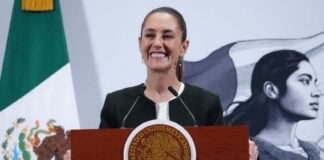President Claudia Sheinbaum standing at the presidential podium at the National Palace with a wide smile as she gazes out at reporters off camera. Behind her is a large Mexican flag on a post and a wall with the logo of Sheinbaum's admnistration: an illustration of a young Indigenous Mexican woman in side profile looking off into the distance, standing in front of the Mexican flag.