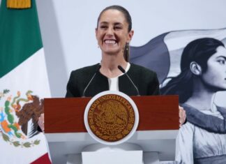 President Claudia Sheinbaum standing at the presidential podium at the National Palace with a wide smile as she gazes out at reporters off camera. Behind her is a large Mexican flag on a post and a wall with the logo of Sheinbaum's admnistration: an illustration of a young Indigenous Mexican woman in side profile looking off into the distance, standing in front of the Mexican flag.