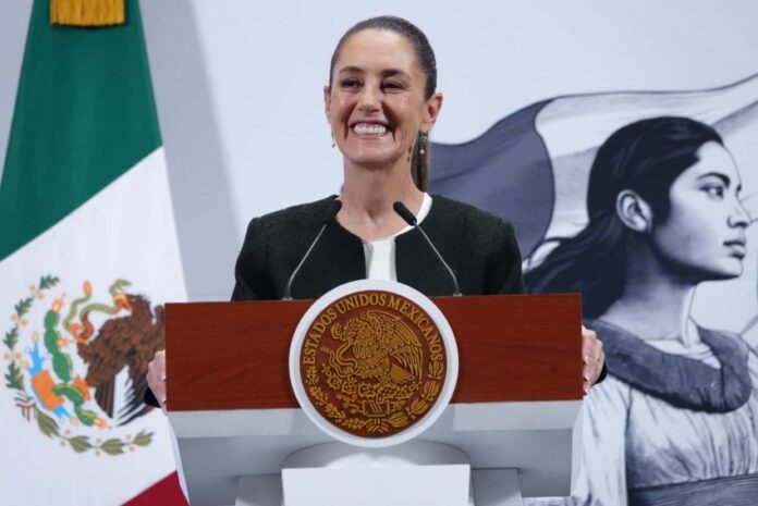 President Claudia Sheinbaum standing at the presidential podium at the National Palace with a wide smile as she gazes out at reporters off camera. Behind her is a large Mexican flag on a post and a wall with the logo of Sheinbaum's admnistration: an illustration of a young Indigenous Mexican woman in side profile looking off into the distance, standing in front of the Mexican flag.