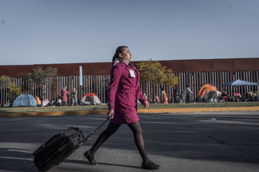 Air steward in a pink skirt suit uniform and an identification tag pinned to it pulls a small black suitcase on wheels behind her as she walks down the street.