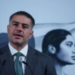 Mexican man in his 40s with a five o'clock shadow and close cropped hair. He's wearing a suit and standing at Mexico's presidential podium with two miniature microphones. Behind him is the black-and-white logo of the current Mexican government, an indigenous Mexican woman in profile, with the Mexican flag behind her.