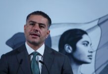 Mexican man in his 40s with a five o'clock shadow and close cropped hair. He's wearing a suit and standing at Mexico's presidential podium with two miniature microphones. Behind him is the black-and-white logo of the current Mexican government, an indigenous Mexican woman in profile, with the Mexican flag behind her.