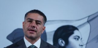 Mexican man in his 40s with a five o'clock shadow and close cropped hair. He's wearing a suit and standing at Mexico's presidential podium with two miniature microphones. Behind him is the black-and-white logo of the current Mexican government, an indigenous Mexican woman in profile, with the Mexican flag behind her.