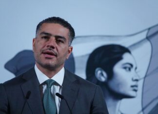 Mexican man in his 40s with a five o'clock shadow and close cropped hair. He's wearing a suit and standing at Mexico's presidential podium with two miniature microphones. Behind him is the black-and-white logo of the current Mexican government, an indigenous Mexican woman in profile, with the Mexican flag behind her.