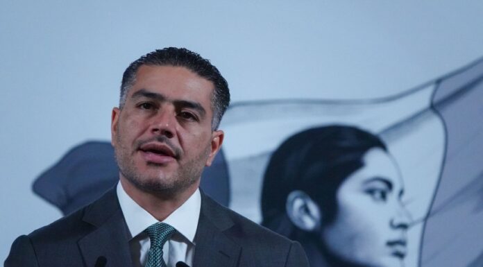 Mexican man in his 40s with a five o'clock shadow and close cropped hair. He's wearing a suit and standing at Mexico's presidential podium with two miniature microphones. Behind him is the black-and-white logo of the current Mexican government, an indigenous Mexican woman in profile, with the Mexican flag behind her.