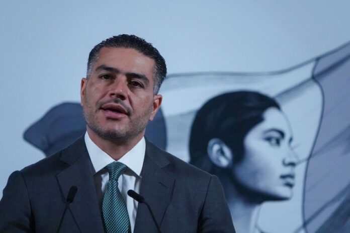 Mexican man in his 40s with a five o'clock shadow and close cropped hair. He's wearing a suit and standing at Mexico's presidential podium with two miniature microphones. Behind him is the black-and-white logo of the current Mexican government, an indigenous Mexican woman in profile, with the Mexican flag behind her.