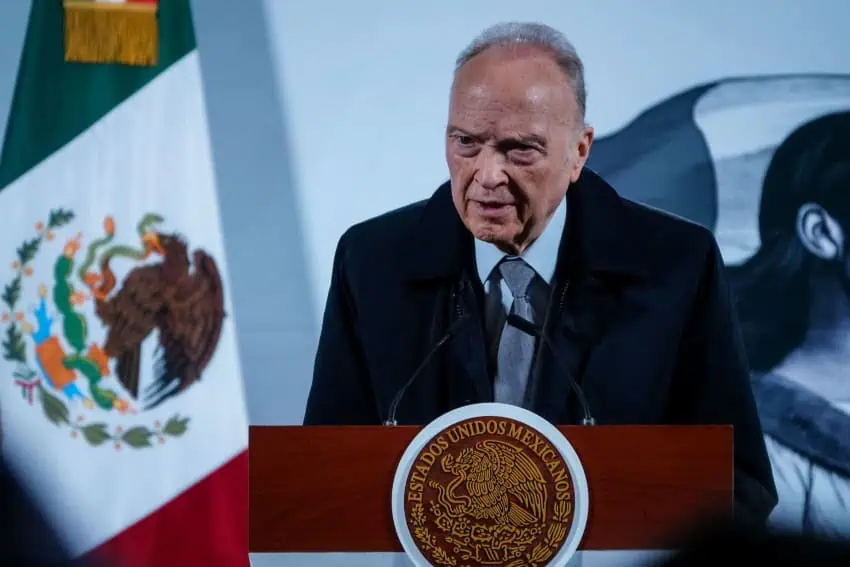 An elderly official in a dark suit and light blue shirt and tie stands at Mexico's presidential podium during a press briefing at the National Palace