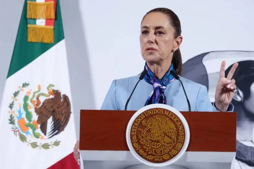 Mexican President Claudia Sheinbaum at the presidential podium in the National Palace, wearing a powder-blue suit jackets and a multicolored scarf around her neck. She is holding up the index and middle fingers of her left hand as she talks to reporters off camera.