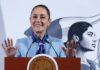 Mexican President Claudia Sheinbaum standing at the presidential podium during her mananera daily press conference. She has both hands raised with palms outward