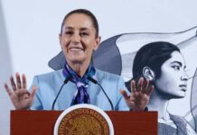 Mexican President Claudia Sheinbaum standing at the presidential podium during her mananera daily press conference. She has both hands raised with palms outward