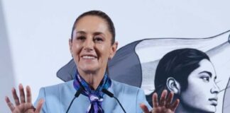 Mexican President Claudia Sheinbaum standing at the presidential podium during her mananera daily press conference. She has both hands raised with palms outward