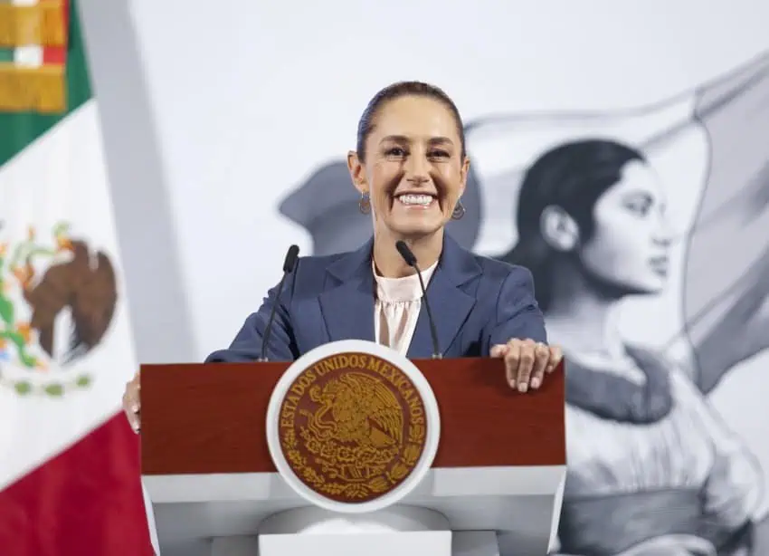 A smiling President Claudia Sheinbaum standing at the presidential podium at Mexico's National Palace as she looks out at reporters off camera.