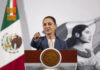 President Claudia Sheinbaum at her daily press conference, standing at the presidential podium in the National Palace in Mexico City. She points at a reporter off camera to take their question.