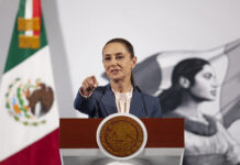 President Claudia Sheinbaum at her daily press conference, standing at the presidential podium in the National Palace in Mexico City. She points at a reporter off camera to take their question.