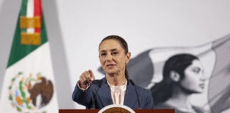 President Claudia Sheinbaum at her daily press conference, standing at the presidential podium in the National Palace in Mexico City. She points at a reporter off camera to take their question.